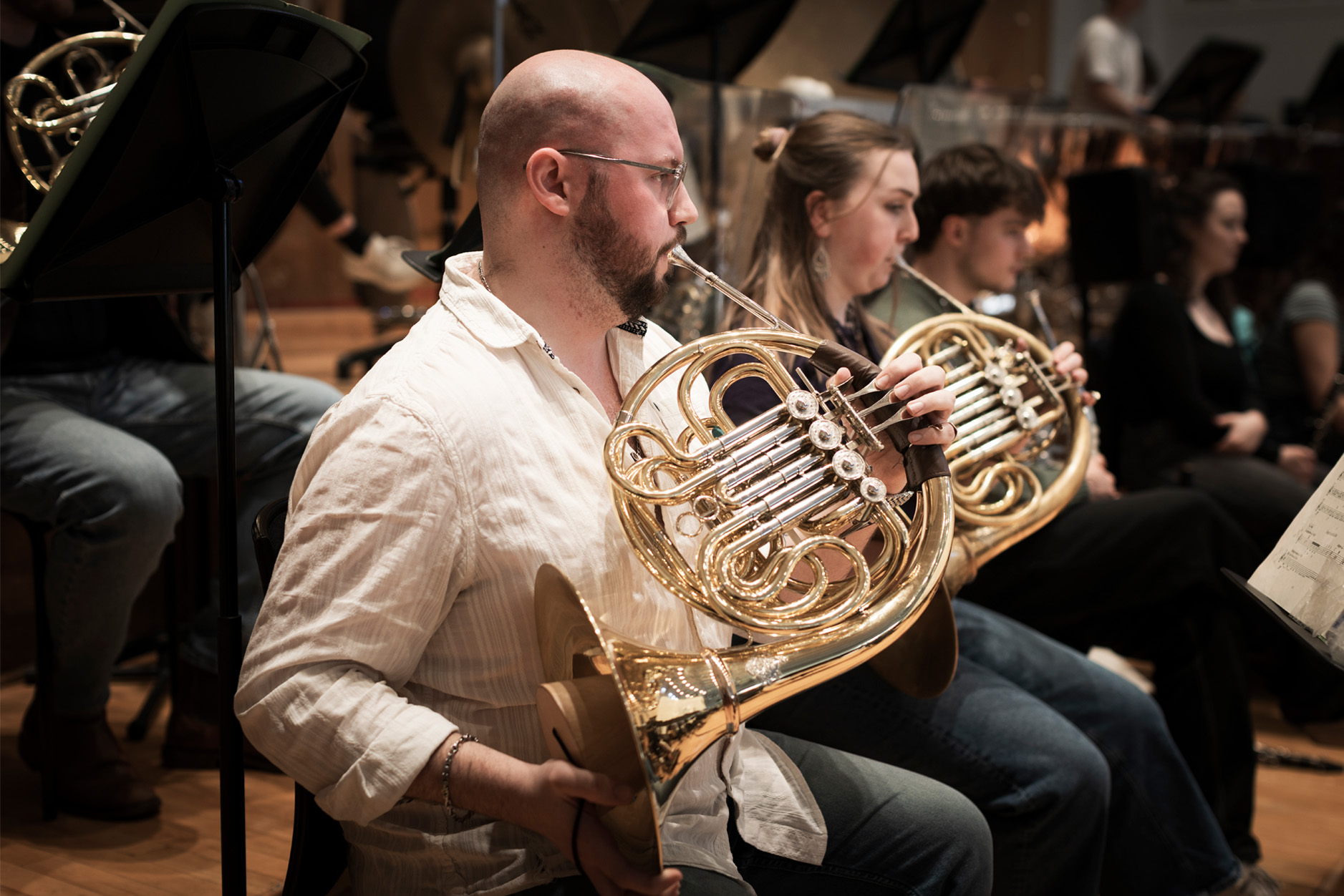 A male and female student, playing the French horns in smart attire in an orchestral performance.